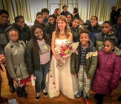 Katya Grineva in a pale floor-length gown holding a bouquet, with children in winter coats standing on either side of her