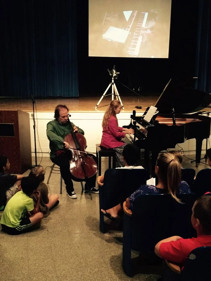 Katya Grineva playing a grand piano beside a seated cellist in a darkened school auditorium, with children sitting on the floor and in seats in front of them