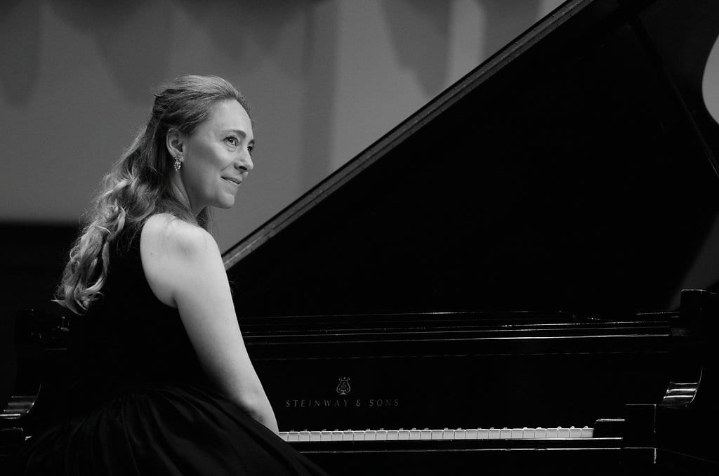 Black and white portrait of Katya Grineva seated at a grand piano, turned toward the camera