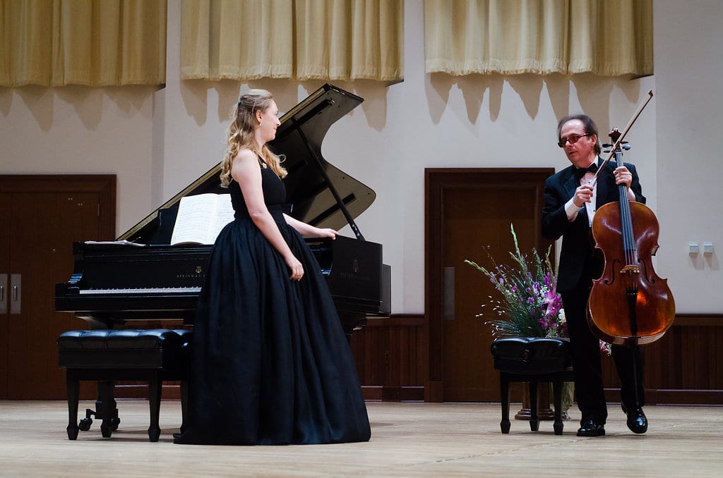 Katya Grineva standing by the piano in a dark gown, a cellist playing at her right