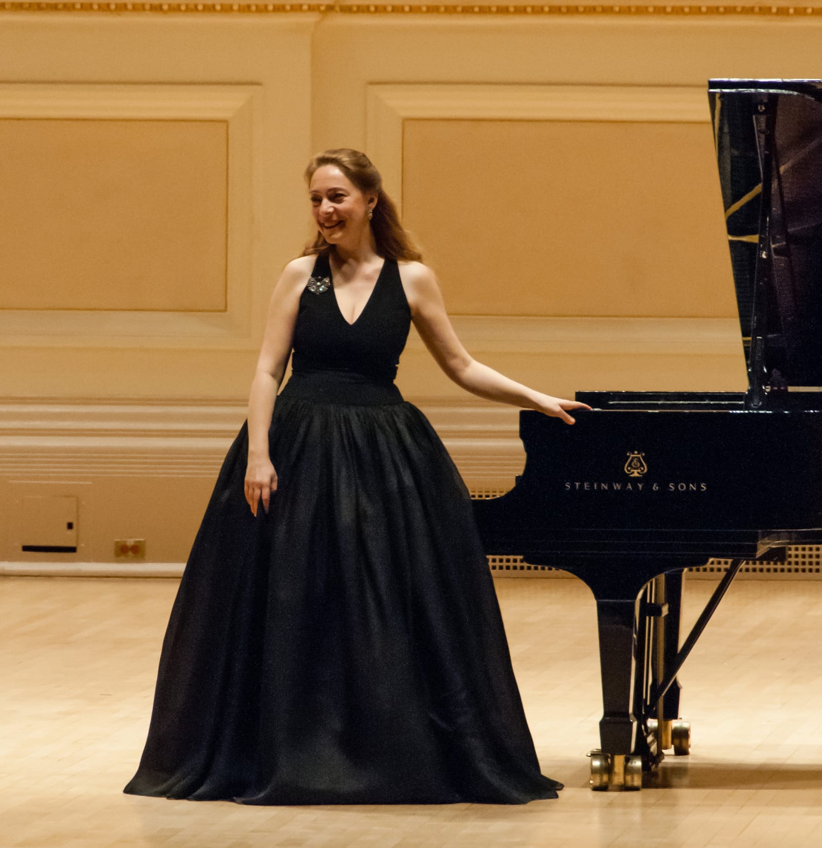 Katya Grineva laughing beside the open Steinway in a black gown
