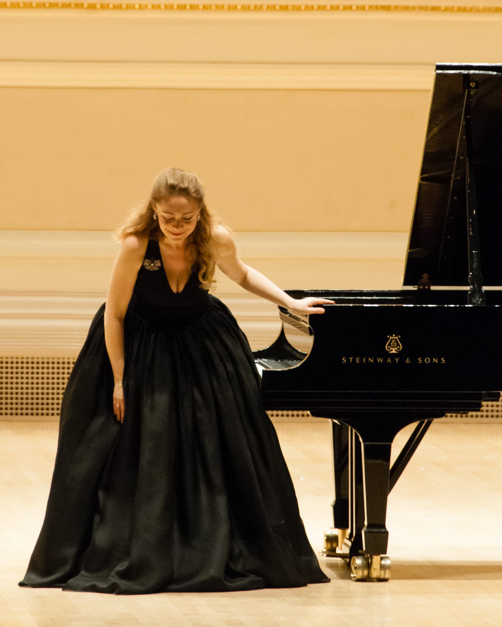 Katya Grineva bowing with one hand resting on the piano lid, in a black gown