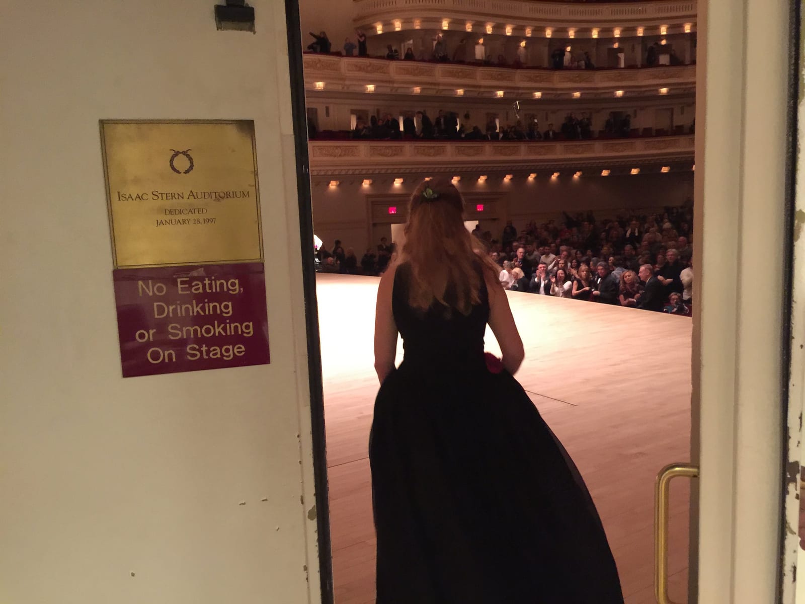 Katya Grineva from behind in a black gown, waiting in the backstage doorway that opens onto the Carnegie Hall stage