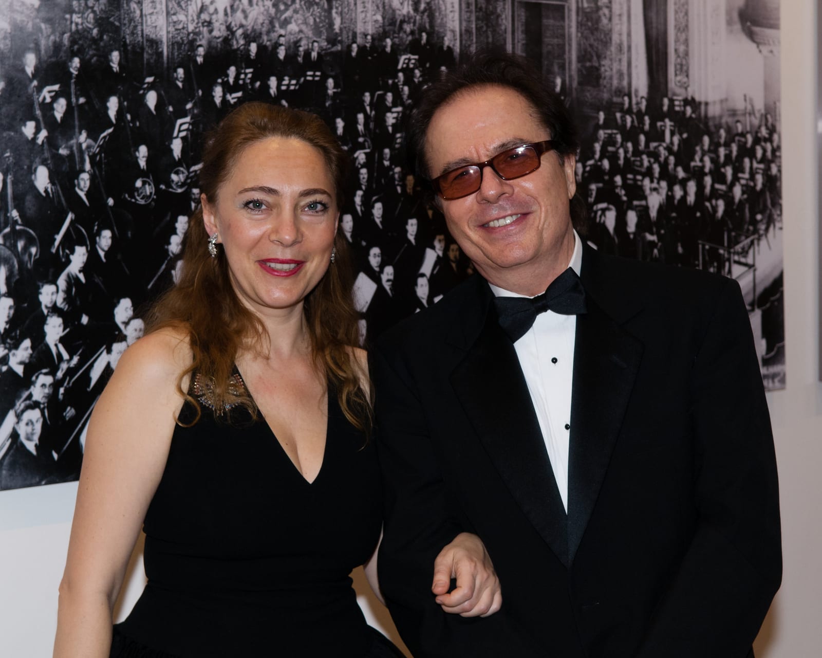 Katya Grineva with a man in a tuxedo and dark glasses, a large black and white orchestra photograph on the wall behind them
