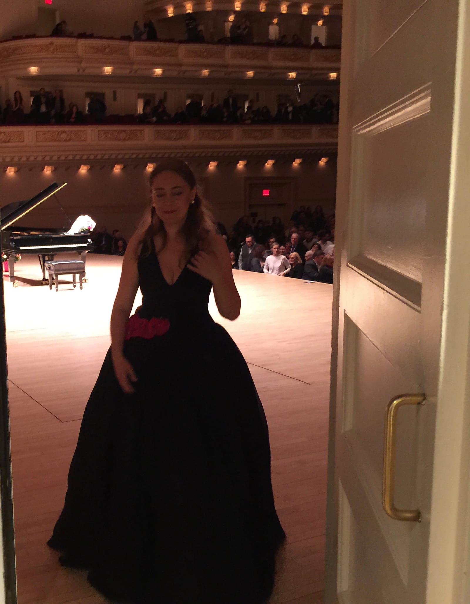 Katya Grineva framed in the backstage doorway, the lit hall filling the space behind her