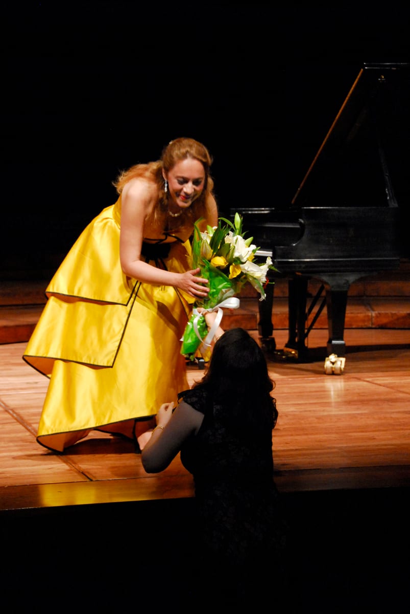 Katya Grineva leaning down from the stage in a yellow gown to take lilies from a kneeling audience member