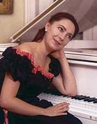 An early portrait of Katya Grineva in a black and red off shoulder dress, leaning on the piano