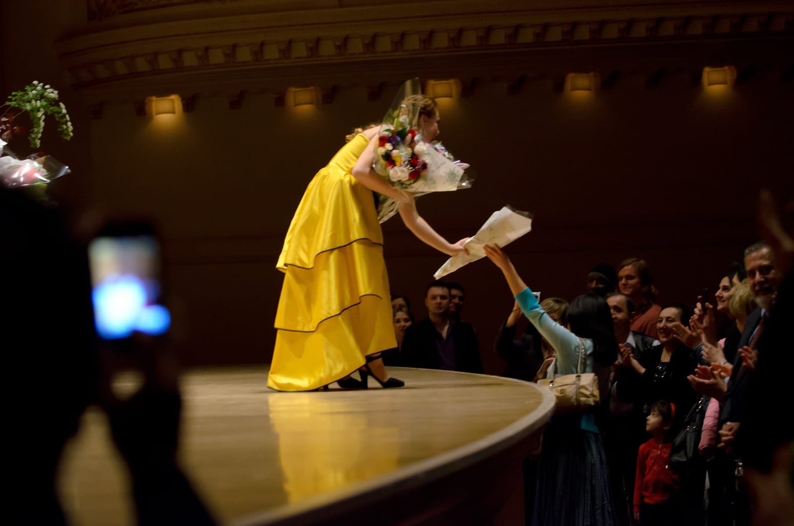 Katya Grineva at the stage edge in the yellow gown, taking flowers from the audience while a phone films from the front row