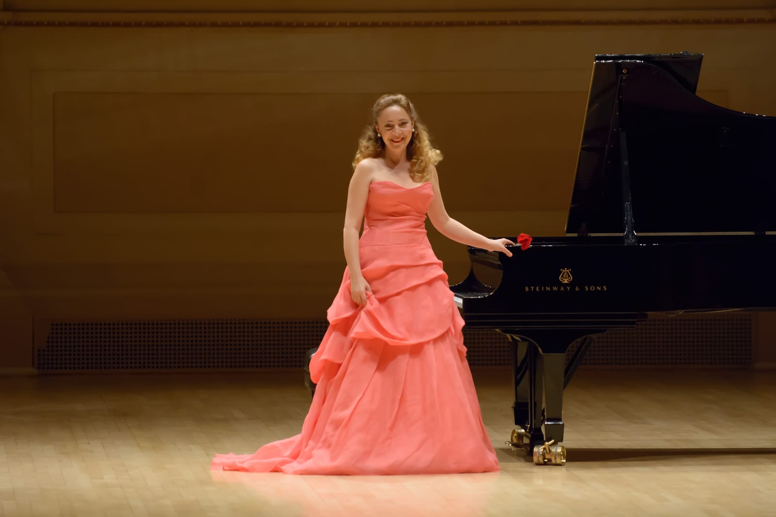 Katya Grineva in a coral gown beside the Steinway, holding a single red rose