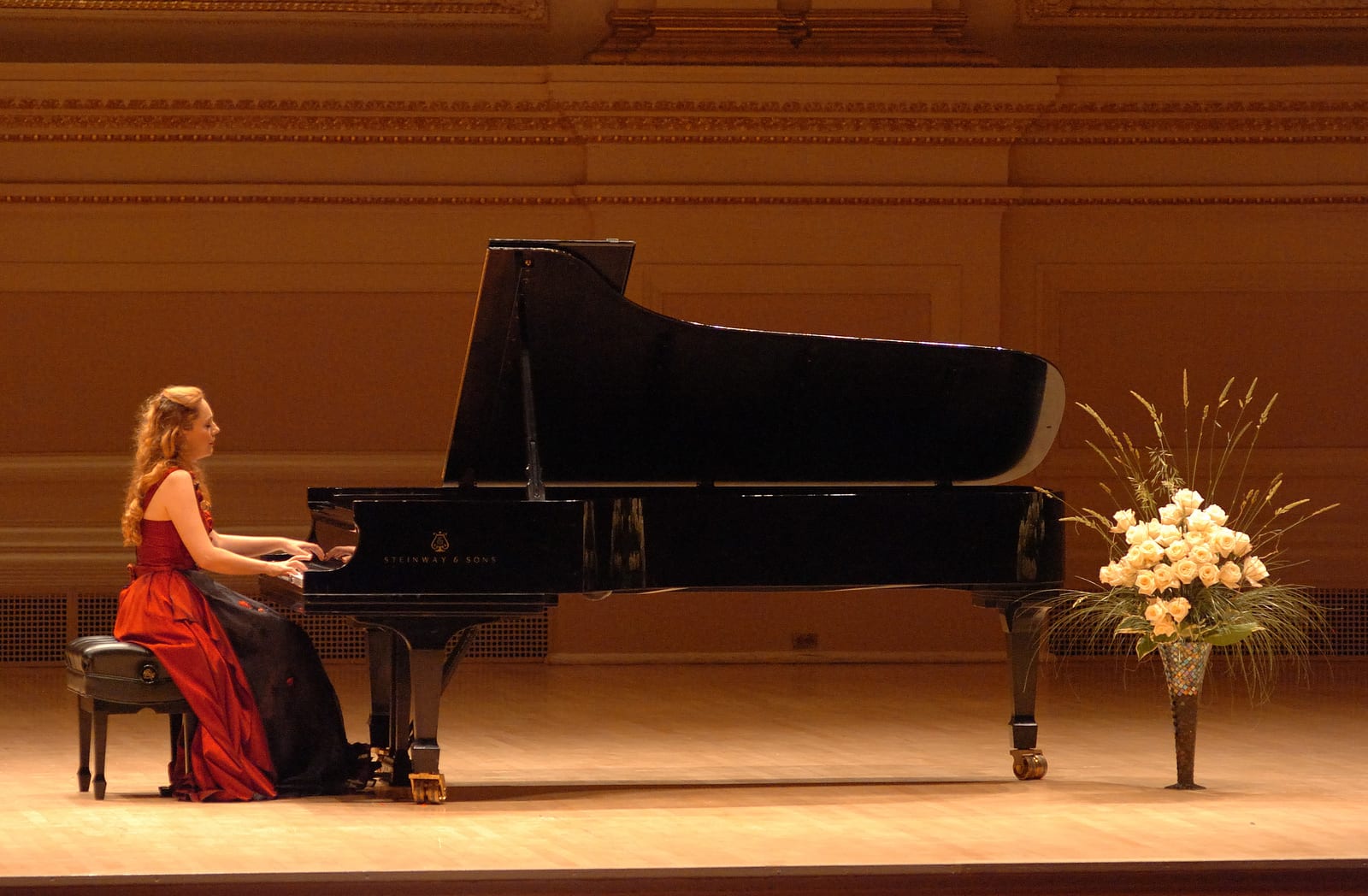 Katya Grineva in a red gown at the piano, a tall white floral arrangement standing at her right