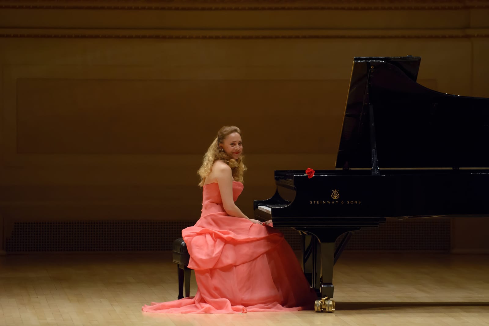 Katya Grineva seated at the Steinway on an empty concert stage in a coral gown, turned toward the camera