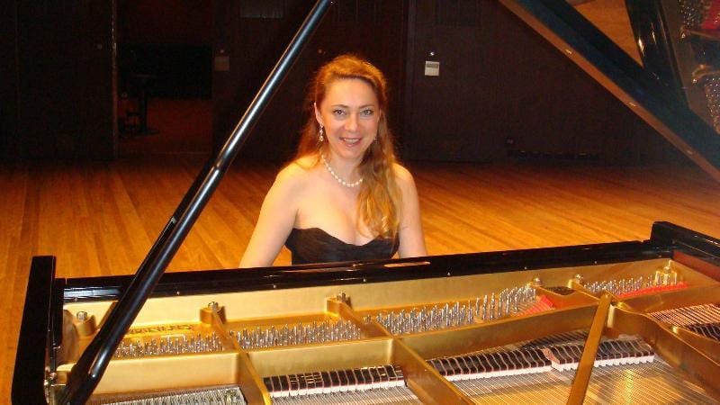 Katya Grineva seated at an open grand piano, smiling toward the camera