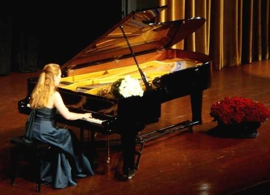 Katya Grineva at an open grand piano on a wooden stage, red flowers banked at her right