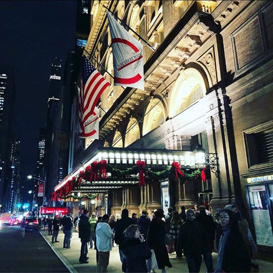 Carnegie Hall at night from 57th Street, an American flag over the lit red awning and people passing on the sidewalk