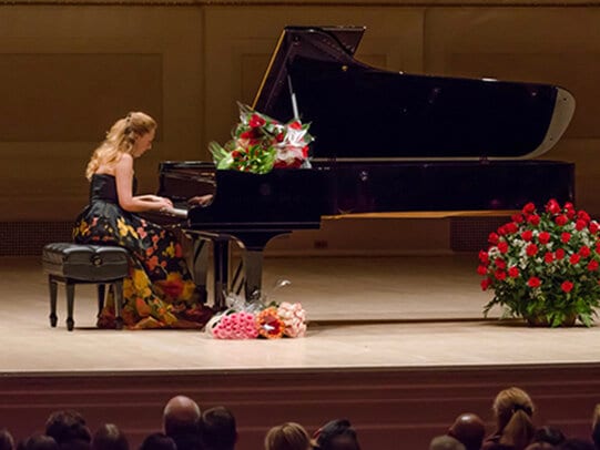 Katya Grineva at the piano in a dark gown, bouquets across the lid and a basket of red roses at her right, the audience in silhouette