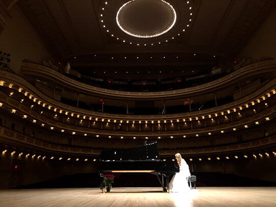 The Carnegie Hall auditorium from stage level, tiers of lit boxes rising into the dark, Katya Grineva small and in white at the piano