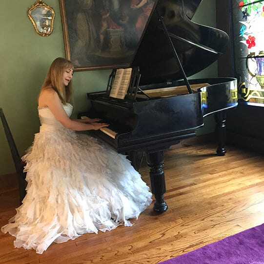 Katya Grineva in an ivory tulle gown at a black grand piano in a private room, an oil painting and stained glass behind her