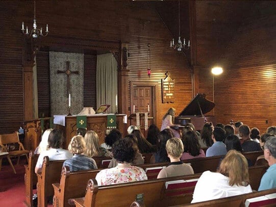 Katya Grineva at a grand piano at the front of a church, the congregation in the pews before her