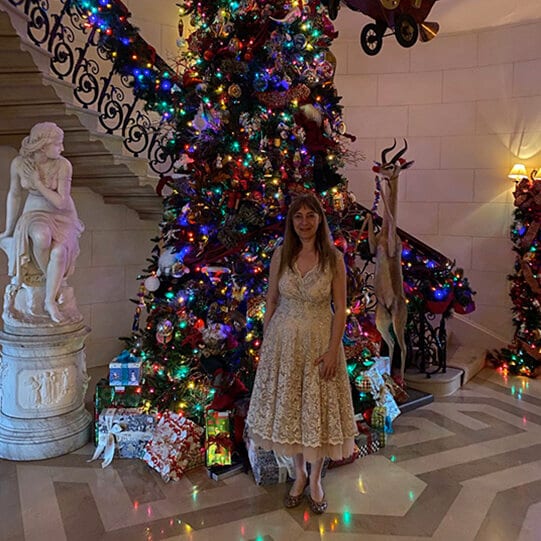 Katya Grineva in a cream lace dress beside a lit Christmas tree, a marble staircase and statue behind her