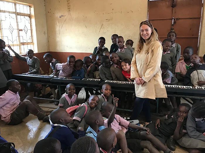 Katya Grineva standing among a large group of schoolchildren in a classroom lined with electric keyboards