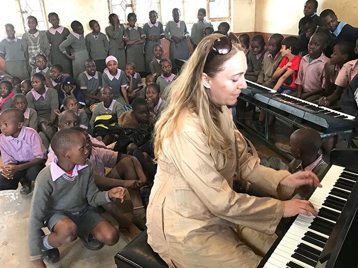 Katya Grineva playing an electric keyboard in a bare classroom, with dozens of children in grey school uniforms seated on the floor and standing close around her