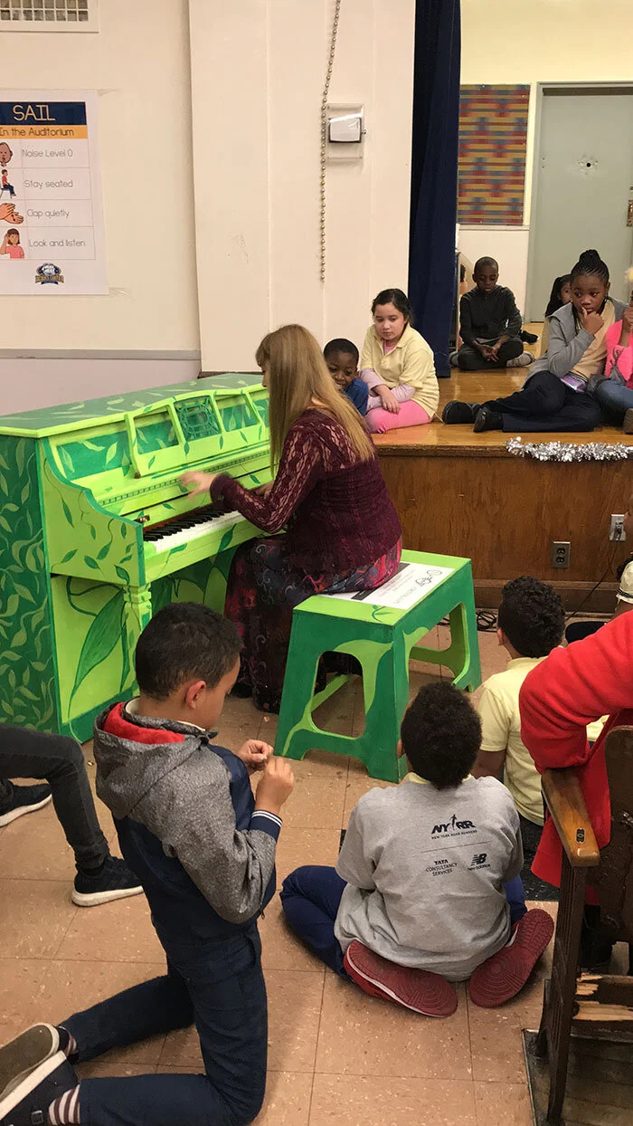 Katya Grineva playing a bright green hand-painted upright piano in a school hall, with children gathered on the floor around her and more seated on the stage behind