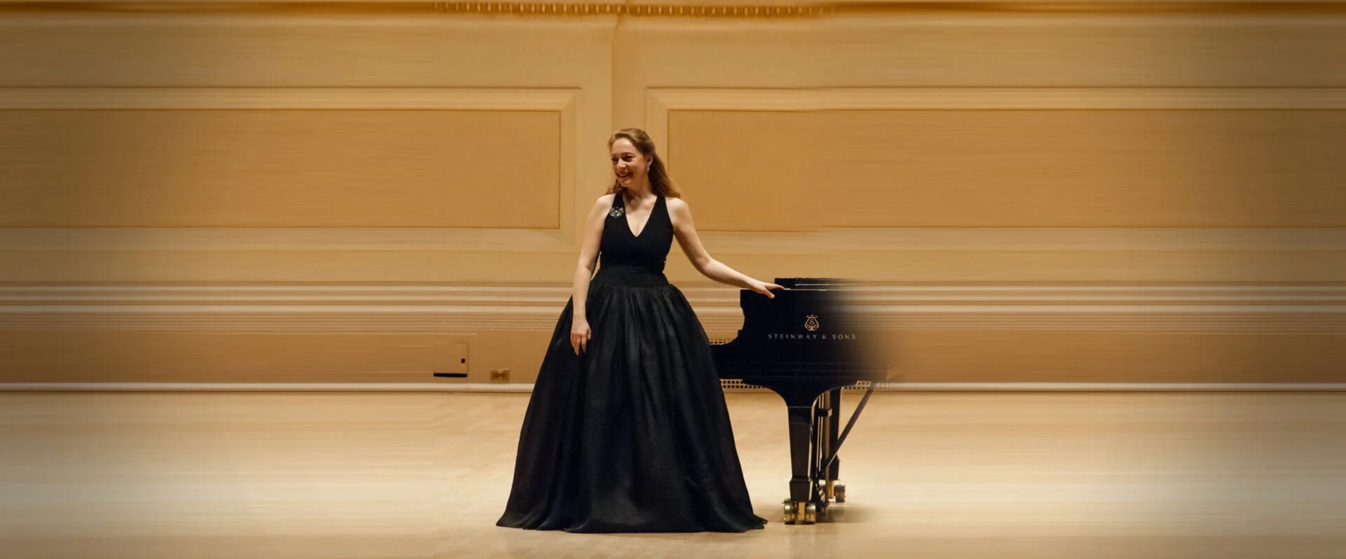 Katya Grineva standing beside a Steinway grand piano on the stage of Carnegie Hall, before an empty hall of warm gold panelling
