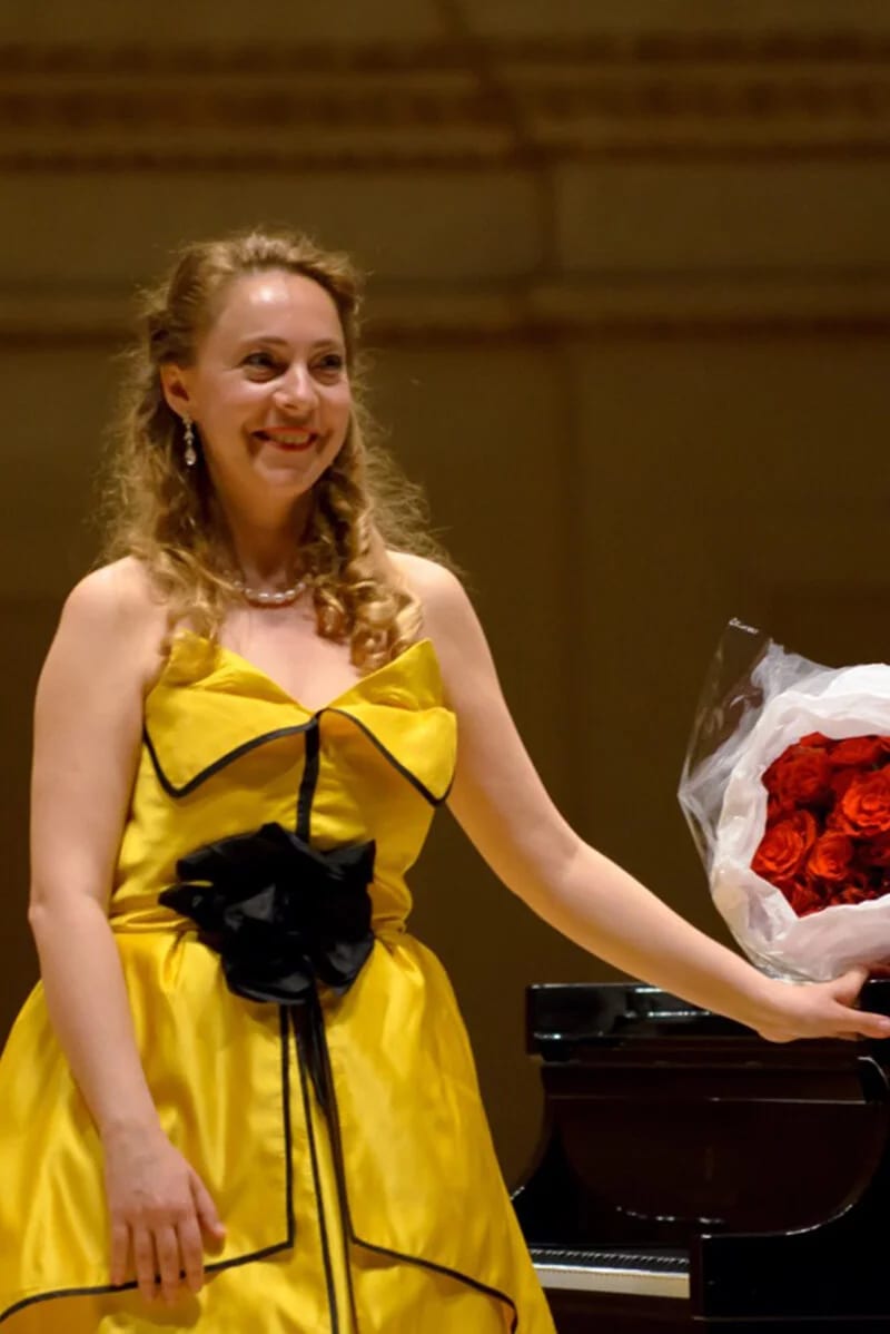 Katya Grineva takes a bow beside the piano, holding a bouquet of red roses