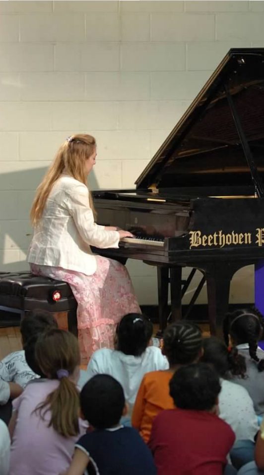Katya Grineva playing a grand piano in a school hall, with children sitting on the floor a few feet in front of her