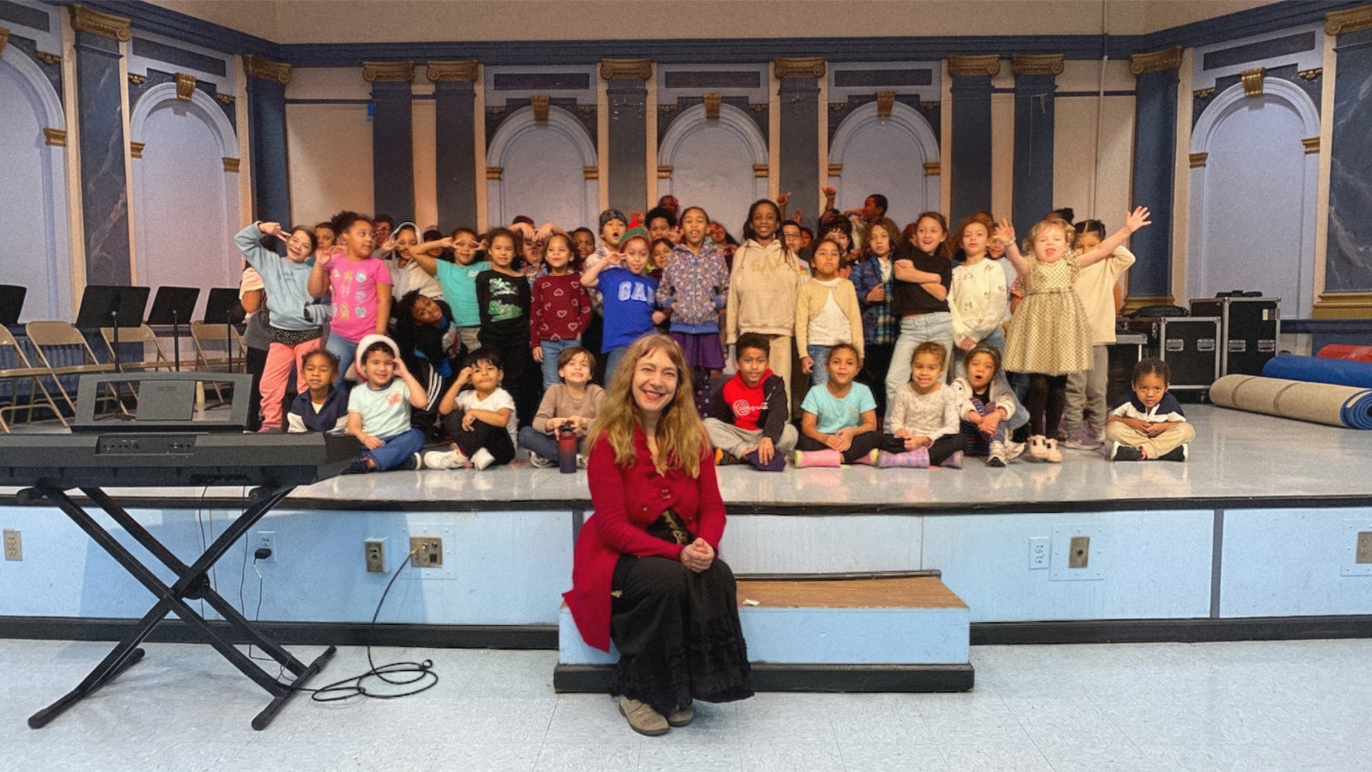 Katya Grineva sits at the front of a school stage in a red cardigan, with about forty children standing and seated on the stage behind her