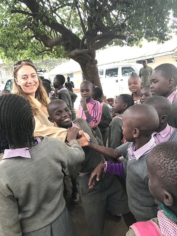 Katya Grineva outdoors under a large tree, greeted by a crowd of schoolchildren in grey uniforms, a laughing boy reaching toward her in the foreground